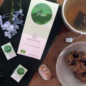 Semper Tea Verde junto a una taza de té, flores y galletas saludables.